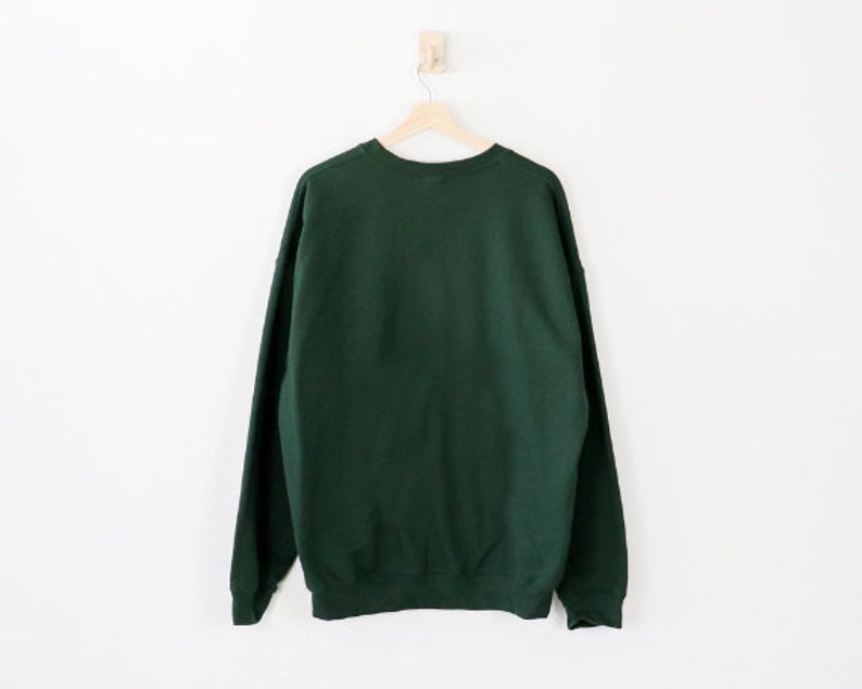 May include: A dark green sweatshirt hangs on a wooden hanger against a white wall. The crew neck sweatshirt is made of a soft material and has long sleeves. The garment is displayed from the back.