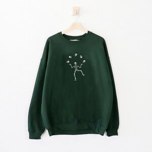 May include: A dark green sweatshirt with a white graphic of a skeleton juggling skulls. The sweatshirt is hanging on a wooden hanger against a white wall. The design is centered on the front of the garment.