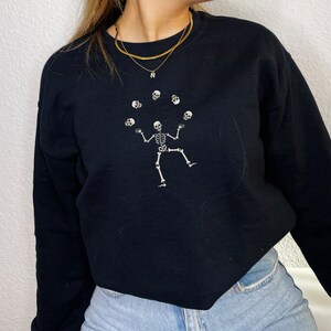 May include: Black sweatshirt with a white skeleton design juggling skulls. The skeleton is in the center, with six skulls above. The sweatshirt has a crew neck and is made of a soft material.