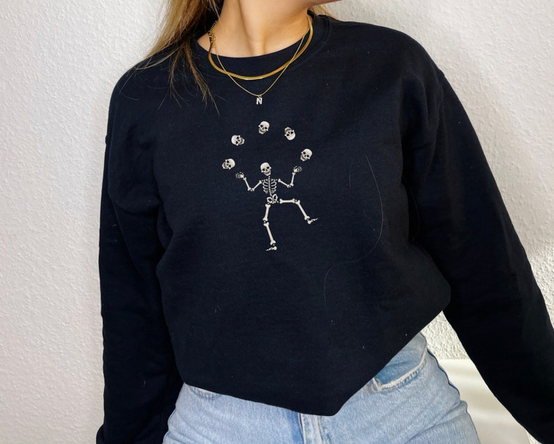 May include: Black sweatshirt with a white skeleton design juggling skulls. The skeleton is in the center, with six skulls above. The sweatshirt has a crew neck and is made of a soft material.