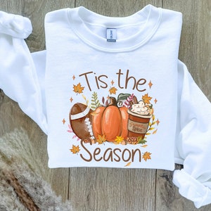 May include: White sweatshirt with a fall-themed graphic. The design includes a football, pumpkins, and a pumpkin spice latte. Text on the shirt reads "Tis the Season" in brown lettering, surrounded by autumn leaves and stars.