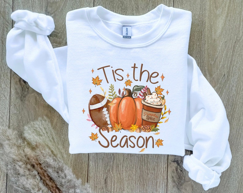 May include: White sweatshirt with a fall-themed graphic. The design includes a football, pumpkins, and a pumpkin spice latte. Text on the shirt reads "Tis the Season" in brown lettering, surrounded by autumn leaves and stars.