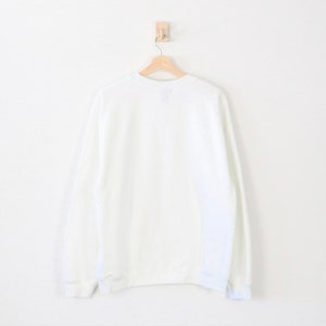 May include: A plain white sweatshirt hangs on a wooden hanger against a white wall. The long-sleeved garment has a crew neck and ribbed cuffs and hem. The sweatshirt is a solid, neutral color.