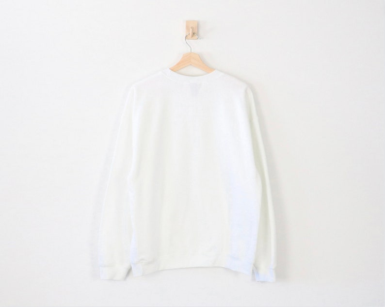 May include: A plain white sweatshirt hangs on a wooden hanger against a white wall. The long-sleeved garment has a crew neck and ribbed cuffs and hem. The sweatshirt is a solid, neutral color.