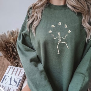 May include: Olive green sweatshirt featuring a skeleton juggling skulls. The graphic is in a cream color. The sweatshirt has a crew neck and long sleeves. The overall style is casual and suitable for fall or Halloween.