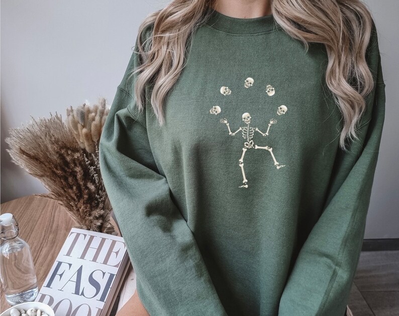May include: Olive green sweatshirt featuring a skeleton juggling skulls. The graphic is in a cream color. The sweatshirt has a crew neck and long sleeves. The overall style is casual and suitable for fall or Halloween.