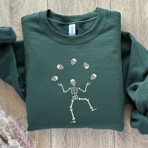 May include: A dark green sweatshirt with a skeleton design juggling skulls. The skeleton is illustrated in a whimsical style, with the skulls suspended above its hands. The sweatshirt has a crew neck and long sleeves.