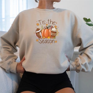 May include: Beige sweatshirt with a fall-themed graphic. The design includes a football, pumpkins, a coffee cup, and the text "Tis the Season" in a decorative font. The sweatshirt is a neutral color, suitable for autumn.