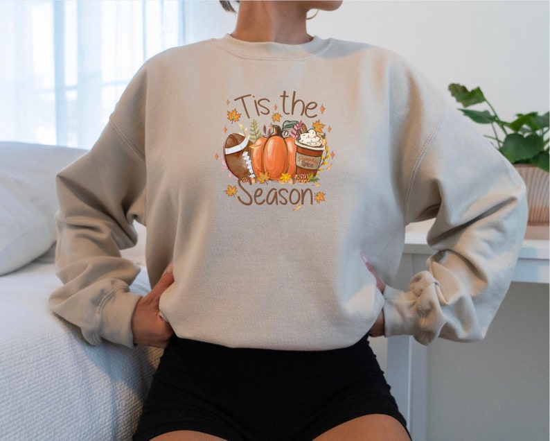 May include: Beige sweatshirt with a fall-themed graphic. The design includes a football, pumpkins, a coffee cup, and the text "Tis the Season" in a decorative font. The sweatshirt is a neutral color, suitable for autumn.