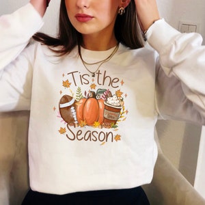 May include: White sweatshirt with a fall-themed graphic. The design includes a football, pumpkins, a coffee cup, and the text "Tis the Season" in brown. Autumn leaves and stars surround the central elements.