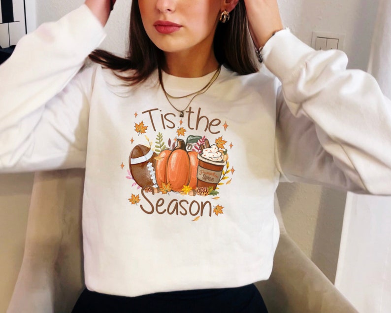 May include: White sweatshirt with a fall-themed graphic. The design includes a football, pumpkins, a coffee cup, and the text "Tis the Season" in brown. Autumn leaves and stars surround the central elements.