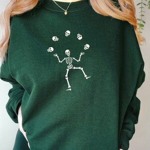 May include: Dark green sweatshirt with a white skeleton juggling skulls. The skeleton is shown with arms outstretched and one leg raised, with six skulls in the air. The sweatshirt has long sleeves and a crew neck.