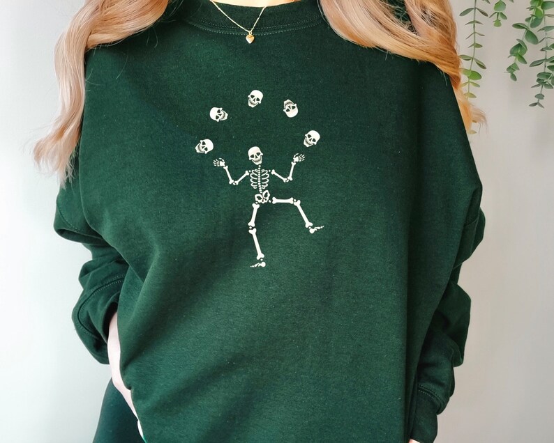 May include: Dark green sweatshirt with a white skeleton juggling skulls. The skeleton is shown with arms outstretched and one leg raised, with six skulls in the air. The sweatshirt has long sleeves and a crew neck.