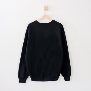 May include: A solid black sweatshirt hangs on a wooden hanger against a white wall. The crew neck sweatshirt has long sleeves and a ribbed hem and cuffs. The garment is made of a soft, comfortable material.