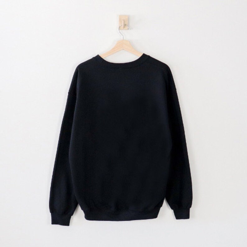 May include: A solid black sweatshirt hangs on a wooden hanger against a white wall. The crew neck sweatshirt has long sleeves and a ribbed hem and cuffs. The garment is made of a soft, comfortable material.