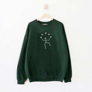 May include: A dark green sweatshirt featuring a white illustration of a skeleton juggling skulls. The garment is suspended on a wooden hanger against a white backdrop. The design is centrally positioned on the front.