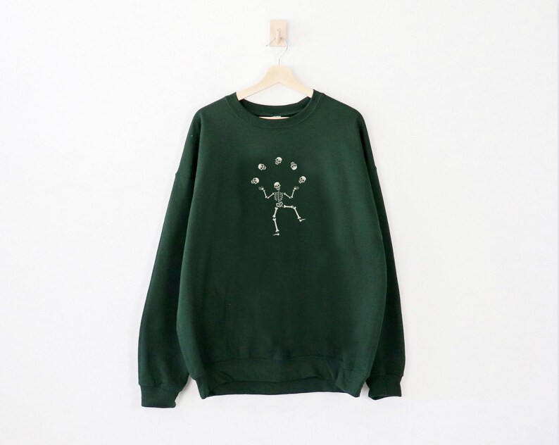 May include: A dark green sweatshirt featuring a white illustration of a skeleton juggling skulls. The garment is suspended on a wooden hanger against a white backdrop. The design is centrally positioned on the front.