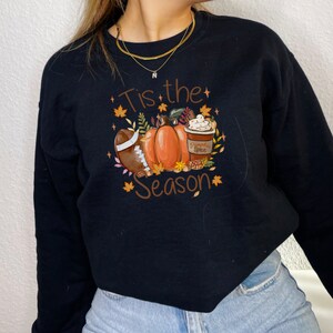 May include: Black sweatshirt with a fall-themed graphic. The design includes a football, pumpkins, a pumpkin spice latte, and the text "Tis the Season" in brown. Autumn leaves and stars surround the central images.