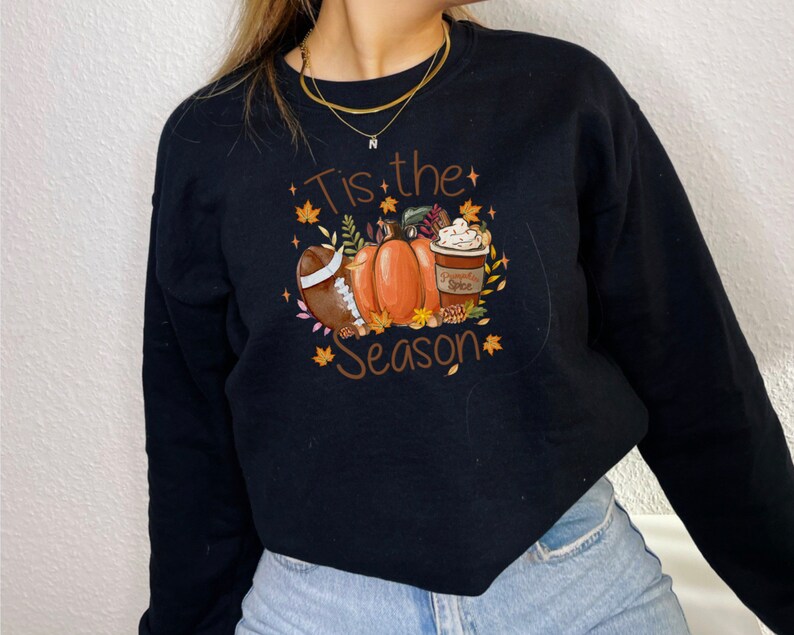 May include: Black sweatshirt with a fall-themed graphic. The design includes a football, pumpkins, a pumpkin spice latte, and the text "Tis the Season" in brown. Autumn leaves and stars surround the central images.
