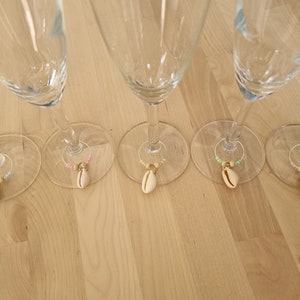 May include: Champagne flutes with seashell wine charms. Each charm has a small seashell pendant, gold-coloured beads, and colourful beads. The charms are attached to silver-coloured rings. The flutes are on a light-coloured wooden surface.