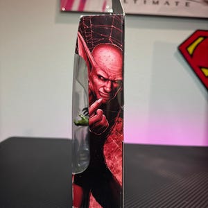 Pode incluir: Vista lateral de uma figura de a&ccedil;&atilde;o embalada com um personagem de pele vermelha com orelhas pontudas e uma express&atilde;o sinistra. A embalagem tem um esquema de cores vermelho e preto com um design de teia de aranha.
