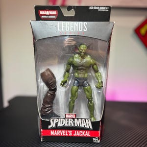 Pode incluir: Uma figura de a&ccedil;&atilde;o Marvel Legends do Jackal da Marvel, um personagem de pele verde, em uma embalagem de pl&aacute;stico transparente. A figura &eacute; articulada e inclui uma bota marrom. A embalagem apresenta o logotipo do Homem-Aranha e o texto "Build-A-Figure".
