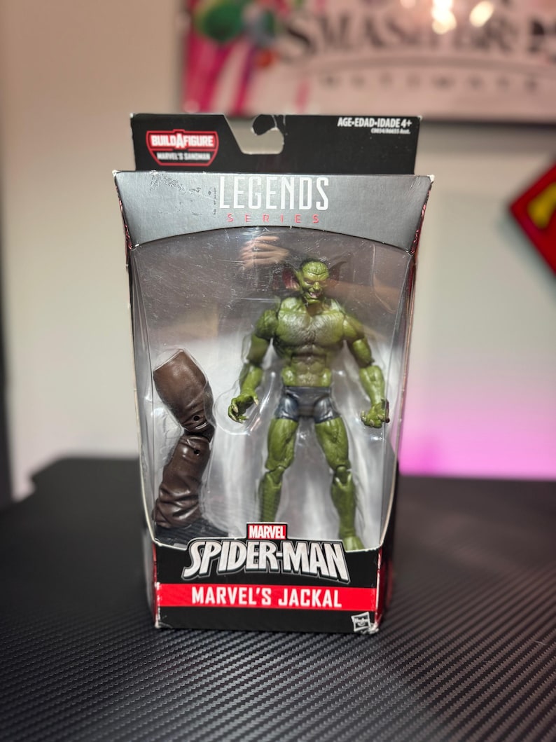 Pode incluir: Uma figura de a&ccedil;&atilde;o Marvel Legends do Jackal da Marvel, um personagem de pele verde, em uma embalagem de pl&aacute;stico transparente. A figura &eacute; articulada e inclui uma bota marrom. A embalagem apresenta o logotipo do Homem-Aranha e o texto "Build-A-Figure".