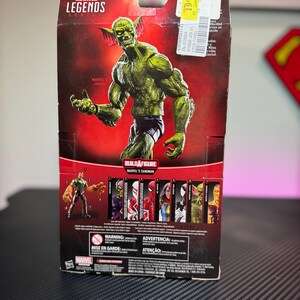 Pode incluir: Uma figura de a&ccedil;&atilde;o Marvel Legends do Jackal, um personagem de pele verde, &eacute; apresentada em uma embalagem do produto. A embalagem inclui o logotipo da Marvel Legends e o texto "Build a Figure Marvel's Sandman". A figura est&aacute; em pose com os bra&ccedil;os estendidos.