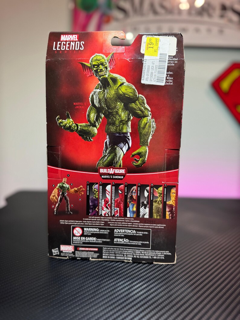Pode incluir: Uma figura de a&ccedil;&atilde;o Marvel Legends do Jackal, um personagem de pele verde, &eacute; apresentada em uma embalagem do produto. A embalagem inclui o logotipo da Marvel Legends e o texto "Build a Figure Marvel's Sandman". A figura est&aacute; em pose com os bra&ccedil;os estendidos.