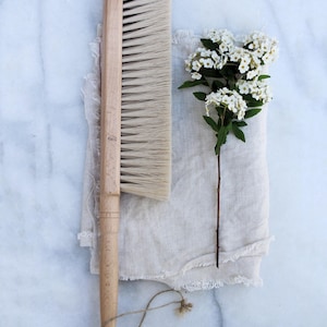May include: A long-handled wooden brush with dense, light-coloured bristles, featuring a small loop of twine at the base of the handle. Beside the brush lies a piece of light beige linen fabric and a single stem of small white flowers.