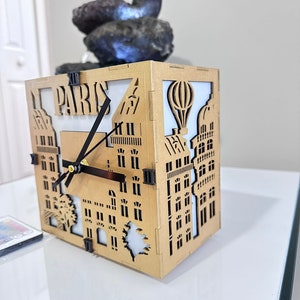 Peut inclure: Une horloge en bois de couleur dorée en forme de bâtiment avec un cadran et des aiguilles noirs. Le cadran de l'horloge porte le mot "PARIS" et un motif de ballon à air chaud.