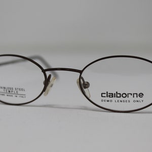 Könnte beinhalten: Braune, ovale Brille mit einem Metallrahmen. Der Rahmen ist mit "Claiborne Demo Lenses Only" und "Stainless Steel Temple Frame Made in Italy" beschriftet.