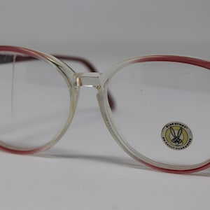 Puede incluir: Gafas de plástico rojo y transparente con un logotipo de Bugs Bunny en la lente derecha. El logotipo dice "Bugs Bunny" y "Not Fault Guarantee".