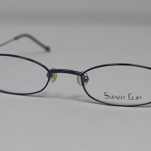 Puede incluir: Un par de gafas de metal gris oscuro con lentes ovalados. El nombre de la marca "SMART CLIP" está impreso en la lente derecha.