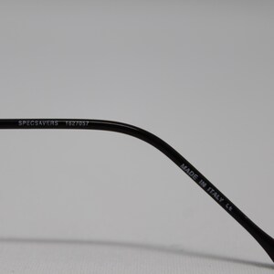May include: Close-up of the black arm of a pair of glasses with the text "SPECSAVERS 1827057" and "MADE IN ITALY CE" printed on it.
