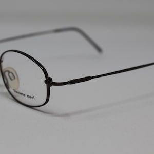 May include: Black metal oval-shaped eyeglasses with clear lenses. The arms of the glasses are thin and have a slight curve. The glasses are made of stainless steel.