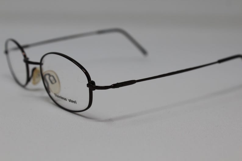May include: Black metal oval-shaped eyeglasses with clear lenses. The arms of the glasses are thin and have a slight curve. The glasses are made of stainless steel.