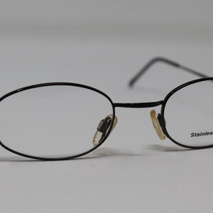 May include: Black oval-shaped eyeglasses with a thin metal frame and clear lenses. The frame is labeled "Stainless steel".