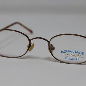 Puede incluir: Gafas de metal ovaladas de color marrón con el texto "ADVANTAGE EYEWEAR" impreso en la lente derecha.