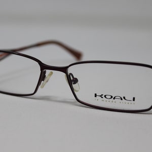 May include: Burgundy-colored metal eyeglasses with a thin frame and a rectangular lens shape. The brand name "KOALI le monde vivant" is printed on the right lens.