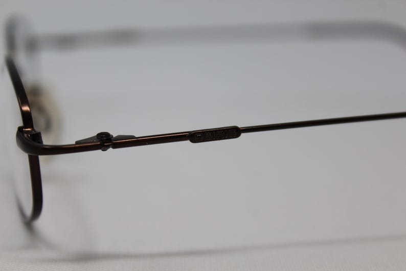 May include: Close-up of the brown metal frame of a pair of eyeglasses. The frame has a spring hinge and the brand name "Calvin Klein" is visible on the arm.