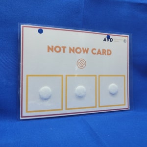 May include: A white card with the text "NOT NOW CARD" in black lettering. The card has three white circles with a red circle with a diagonal line through it in the center. The card is made of thick cardstock and has a clear plastic sleeve.