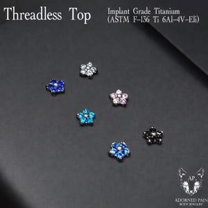 May include: Six flower-shaped threadless tops made of implant grade titanium (ASTM F-136 Ti 6Al-4V-Eli). The jewelry pieces feature various colored gemstones, including blue, clear, pink, and black, arranged in a floral pattern with a central stone.