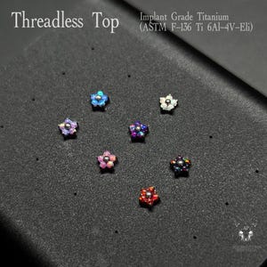 May include: A collection of threadless top body jewelry in a variety of colors, including blue, purple, pink, and red. Each piece is shaped like a five-petal flower. The text "Threadless Top" and "Implant Grade Titanium" are visible.