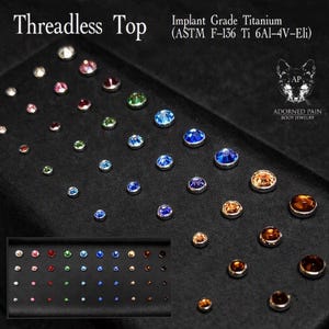 May include: A collection of threadless tops made of implant grade titanium, ASTM F-136 Ti 6Al-4V-Eli. The jewelry pieces feature various colored gemstones in different sizes, arranged on a black display tray with a logo of a cat.