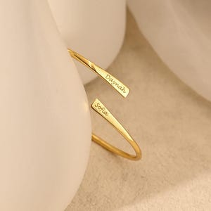 Puede incluir: Una pulsera de brazalete abierta de color dorado con los nombres "Daymah" y "Sofia" grabados. La pulsera se exhibe cerca de jarrones de cerámica blancos y tela blanca.