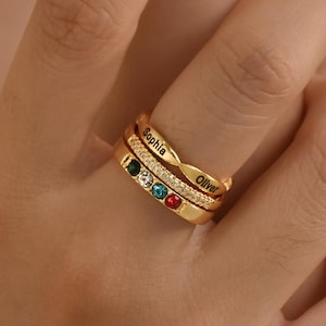 May include: A close-up of gold-coloured rings stacked on a finger. The top ring is engraved with the name "Sophia". The middle ring has a row of clear stones. The bottom ring is engraved with the name "Oliver". The middle ring has three coloured stones: green, blue, and red.