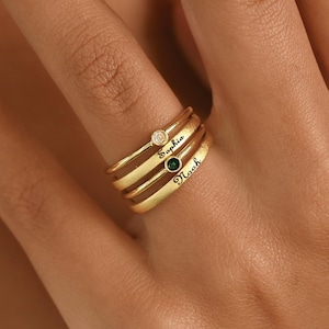 May include: Three gold rings stacked on a finger. Each ring has a different design: a small diamond, a green gemstone, and the name "Noah" engraved. The rings are a stylish accessory.