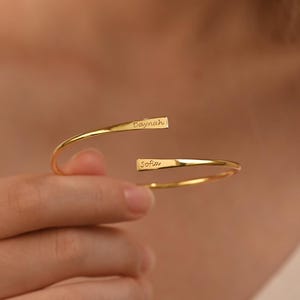 Peut inclure: Un bracelet manchette ouvert de couleur or, avec les noms "Edaynah" et "Sofia" gravés aux extrémités. Le bracelet a un design simple et élégant et est porté au poignet.