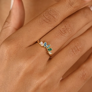 May include: A gold ring featuring a series of colorful, marquise-cut gemstones. The gemstones include light pink, blue, green, and white. The ring is worn on a person's finger.